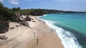 Touristic beach with umbrellas and sunbeds surrounded by cliffs  and azure Indian Ocean with frothy waves on bright sunny day. Aerial footage of Dreamland beach, Bali, Indonesia.