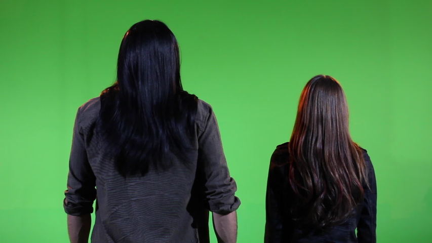 Medium wide 2-shot of two long haired, people standing with backs toward camera as they watch something causing Warm light to pulse on them. Shot in front of green screen.