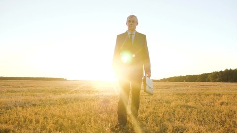 businessman walks through the park in sun. farmer walks across the field. business man with black briefcase walking through field in rays of the bright sun.
