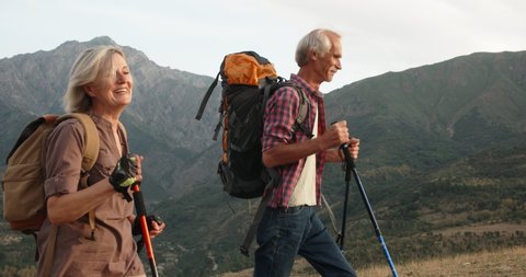 Positive mature caucasian couple trekking in mountains with backpacks, enjoying their adventure 4k