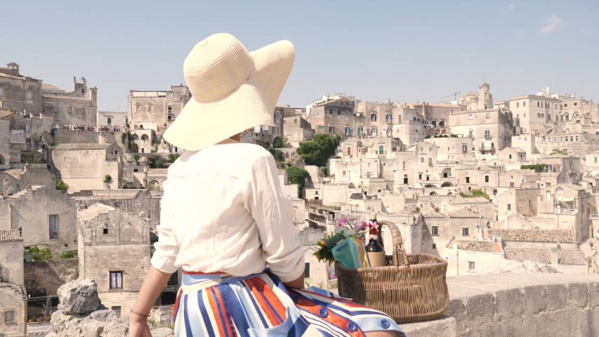 Beautiful elegant young woman in Matera Italy sitting on a high balcony wall viewpoint with landscape. Fashion dress with hat and skirt. 