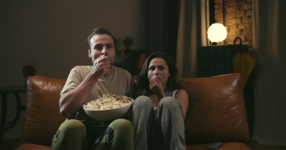 Scared caucasian couple watches movie on couch and eats popcorn at home. They spill popcorn because of unexpected scene Royalty-Free Stock Footage #1030305527