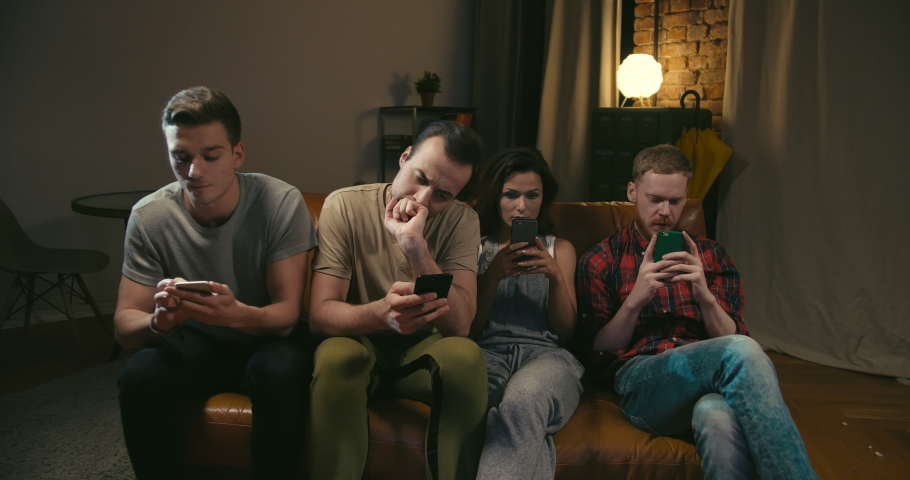 One female and three male caucasian friends are bored, scroll and text with their smartphones during commercial on TV at night in dark room. Terrible party | Shutterstock HD Video #1030305800