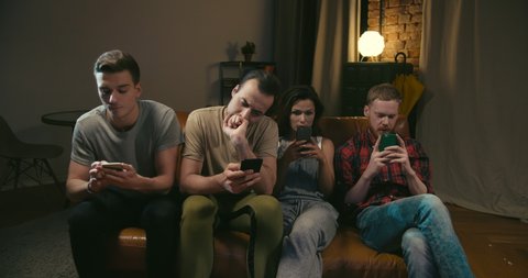 One female and three male caucasian friends are bored, scroll and text with their smartphones during commercial on TV at night in dark room. Terrible party