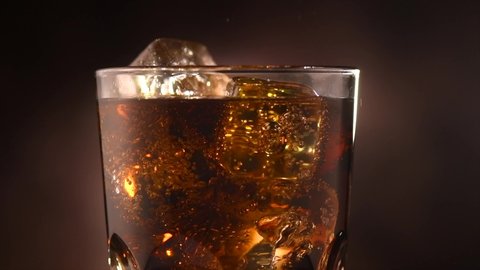 Cola with ice cubes close-up. Cola with Ice and bubbles in glass. Soda closeup. Food background. Rotate glass of Cola fizzy drink over brown background. Slow motion 4K UHD video footage
