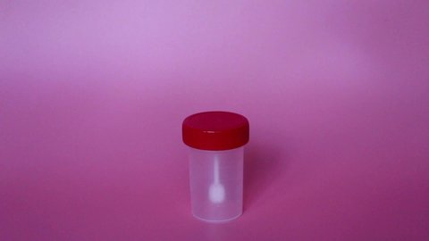 A hand in a blue latex glove shows a container for collecting feces. Pink background. The analysis jar is empty. Medical tests.