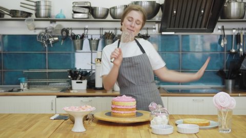 Joyful female pastry chef having fun singing with whisk as microphone and dancing while decorating handmade cake in bakery kitchen, tracking left shot