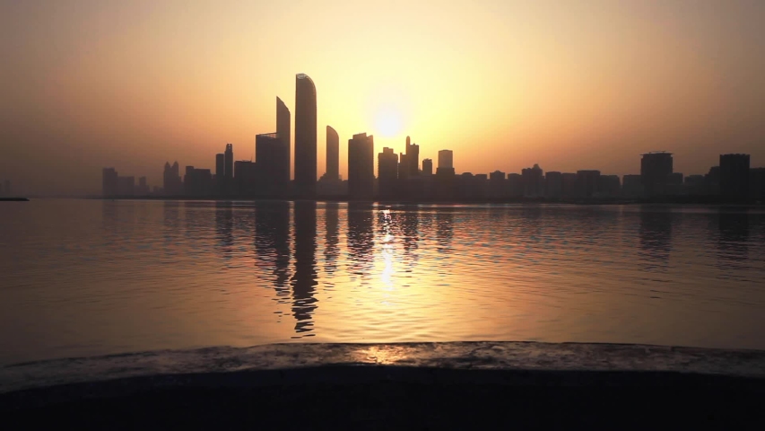 Woman in a long skirt walking across the camera at sunrise. Cityscape of Abu Dhabi at the background.