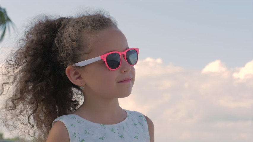 Close-up Portrait of a Beautiful Little Girl in Pink Glasses, Cute Smiling, Looking at the Blue Sky. Concept: Children, Childhood, Summer, Baby, Toddler.
