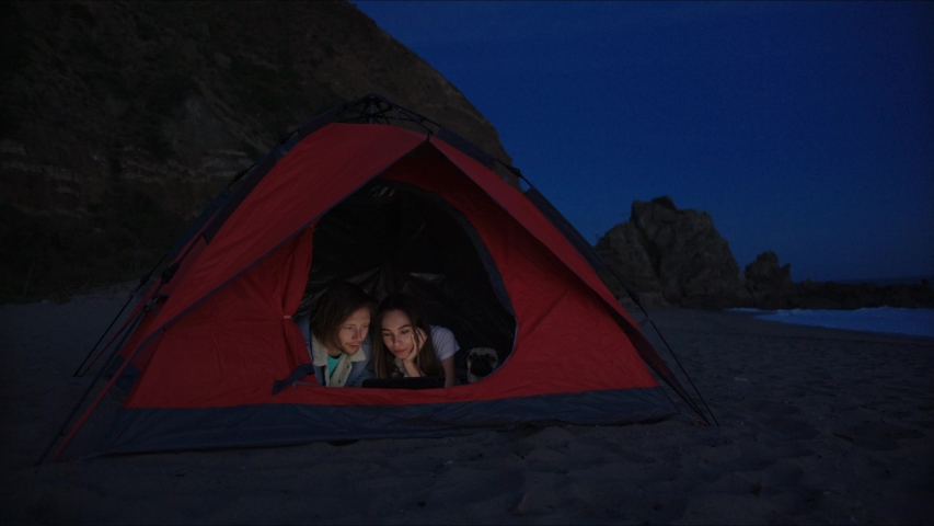 Lovely couple (and dog) watching tv shows in tent using a tablet computer while being on the beach in Malibu at twilight. Camping 