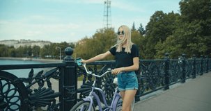 Young woman walking on city streets with bicycle and drinking coffee to go, sunshine, 4K slow motion video footage