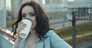 Pretty young woman drinking hot coffee from paper cup. Girl standing on city bridge in summer sunset time and enjoying view, close-up. Urban concept . 4k slow motion video footage 60 fps