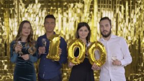 The number 100 of foil. 4 happy cheerful people at a party with balloons in their hands. A group of laughing people of mixed race on a gold background. 4K - Powered by Shutterstock - Get 15% off with code: PIKWIZARD15