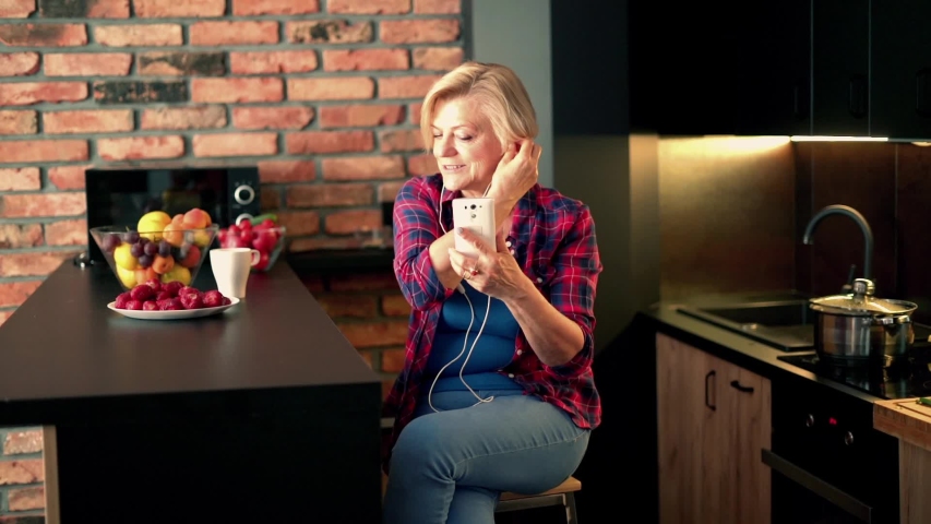Senior, happy woman listening to music on cellphone by table in the kitchen
