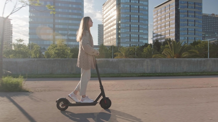 Young beautiful woman riding an electric scooter to work, modern girl, new generation, electric transport, ecology, ecological transport, sunset, electric skateboard, 4k | Shutterstock HD Video #1030375331