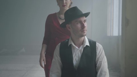 Handsome confident mafia boss in a hat and a vest sitting in an abandoned building. The elegant woman in red dress coming from behind and hugging the man smiling. Happy couple of gangsters