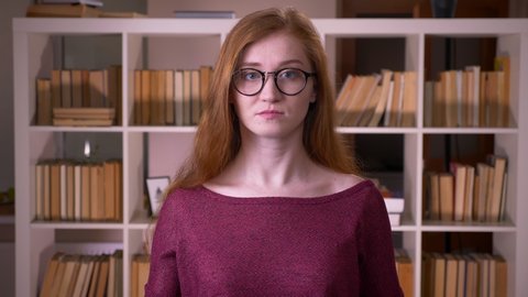 Closeup portrait of young redhead attractive caucasian female student in glasses having her fingers crossed with anxiety looking at camera in the college library