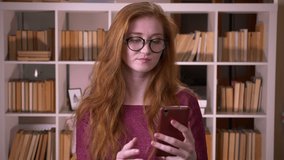 Closeup portrait of young redhead attractive caucasian female student in glasses having a video call on the phone in the college library