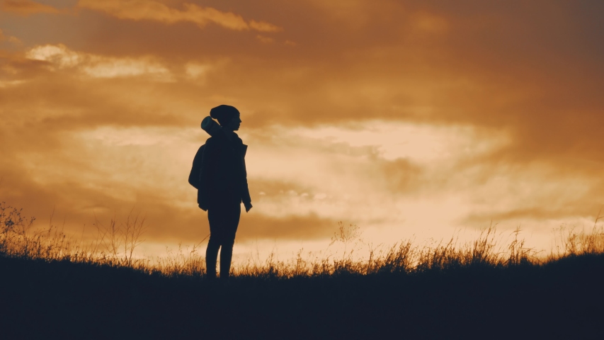 Silhouette of cheerful girl tourist with backpack dancing at sunset, standing on top of a mountain. The concept of active recreation, healthy lifestyle.