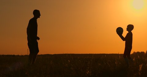 Father and son playing football in the park at sunset, silhouettes against the backdrop of a bright sun, slow-motion shooting