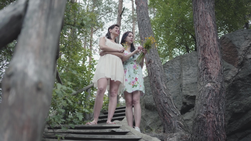 Two young women in short dreses with flowers standing on the stairs and looking at amzing forst, trees, plants. Pretty girls walk outside the city. Cute girlfriends spending weekend together outdoors.