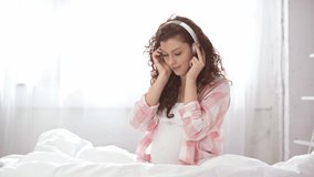 beautiful pregnant young woman listening music, smiling and holding headphones near tummy in bed