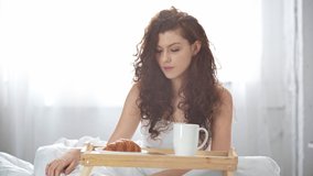 beautiful girl using smartphone, drinking coffee, smiling and looking at camera in bed