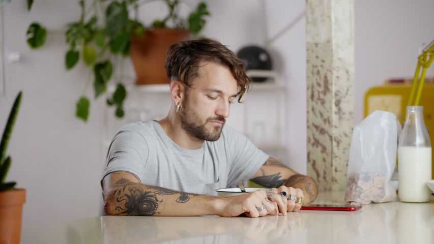 Sleepy tired guy preparing breakfast flakes with milk and using red smartphone on morning, carelessly touching curly messy hair with tattooed arm and joyfully eating sitting at kitchen bar table
