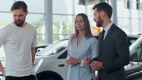 Professional car salesman is telling interested buyers beautiful couple about luxurious car in motor show while man and woman are looking at auto and listening to dealer.