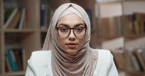 Portrait of female muslim student, wearing hijab, who is at campus library and confidently looking at camera - modern islam concept 4k