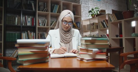 Muslim student is doing research at desk full of books in library. Girl wearing hijab is preparing for exam or making a project - student lifestyle, modern islam close up 4k