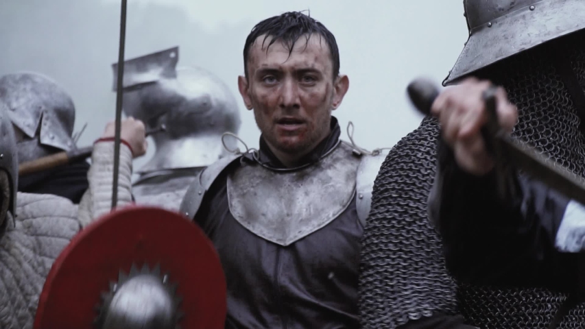 A knight in armor is standing on the battlefield while people are fighting