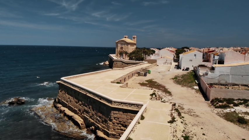 The promenade and the church on the edge of the dreamy village on the small Spanish island Tabarca off the coast of Alicante. There are many seagulls in the air. Sunshine and blue sea. Aerial 4k.