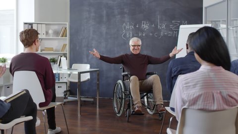 Cheerful mature professor sitting in wheelchair entering classroom and smiling, multiethnic students clapping hands and greeting him