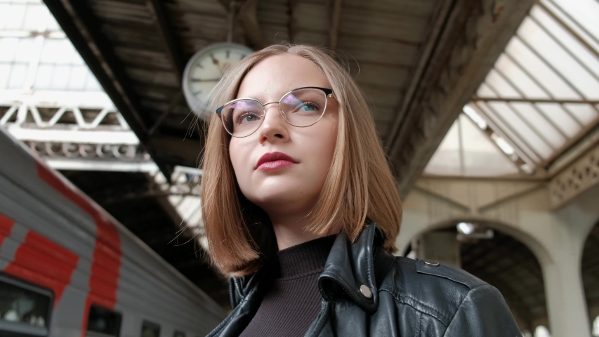 Attractive red-haired young woman with short haircut, wearing glasses and black leather jacket is standing at Vitebsk railway station in St. Petersburg and waiting for train. Big clock.