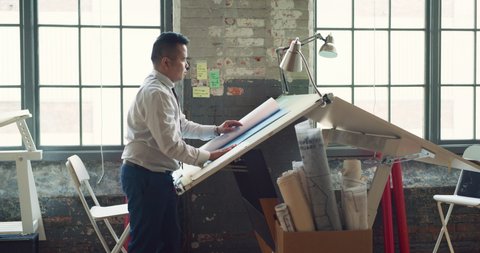 Asian architect working at drafting table drawing architectural sketches in a modern, industrial office and workspace. Business and design thinking. 