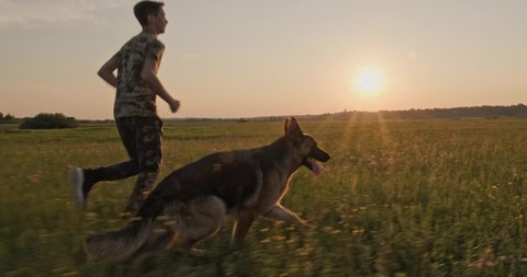 Teenager is running with a dog in nature while sunset. 15 year old boy runs with a dog breed German shepherd. Teen on the field with dog. Caucasian guy is playing with his pet in the meadow. Slow moti