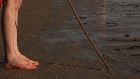 Shot starts close on a female's feet wearing sandals, as she draws in the sand with a stick. The shot then tilts up to reveal a man walking towards her on the beach.