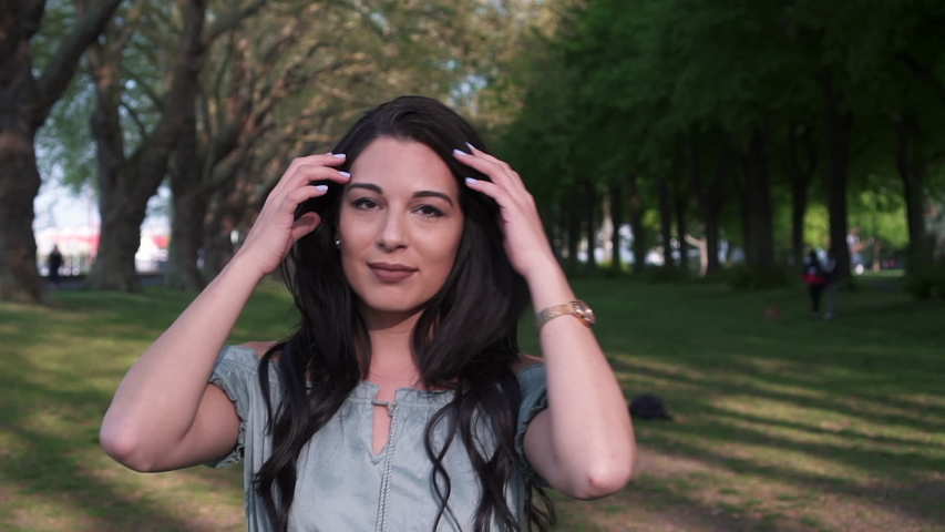 Attractive and playful latina woman with black wavy hair walking under the trees in a park in London, looking at the camera, happy with a beautiful smile.