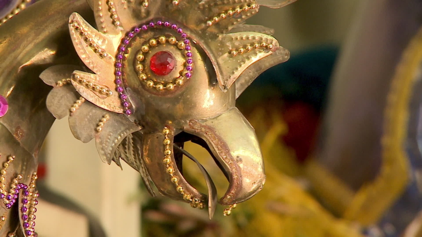 Extreme close-up, low angle still shot of a decorated golden antique head of a tinsmith made rooster with red beads on its eye, surrounded by matching gold and purple beads, Oruro workshop, 
