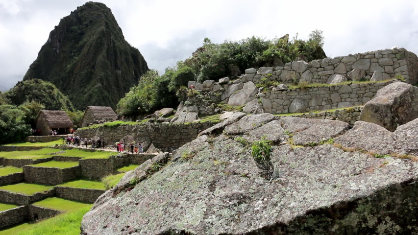 Medium low-angle still shot of a large slanting rock at the top of Machu Picchu ruins, and tourists walking along a horizontal path, Machu Picchu, Peru