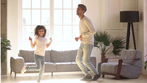 Happy family dad and kid daughter laughing jumping dancing in living room lit with sunlight, energetic child girl having fun with father moving to music play with parent at home enjoy funny activity