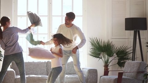 Happy young family mom dad and little kid daughter having fun pillow fight in living room, cheerful mother father child girl laughing feel joy play funny game at home enjoy leisure activity together