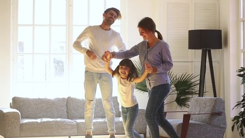 Happy active funny parents and cute little child daughter laughing jumping dancing in modern new house living room, mom dad with kid girl enjoying playing having fun together, family mortgage concept