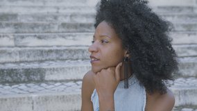 Side view of pretty young Afro-american woman with curly hair and in jeans sleeveless dress sitting outside, looking aside. Lifestyle concept