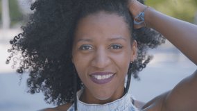 Pretty young Afro-american woman with curly hair and in jeans sleeveless dress standing outside, looking at camera, playing with hair, enjoying her body, sending kisses. Lifestyle concept