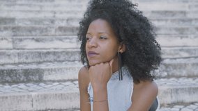 Pretty young Afro-american woman with curly hair and in jeans sleeveless dress sitting outside, looking aside, turning head to camera, smiling. Lifestyle concept