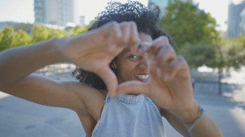 Cheerful young Afro-american woman with curly hair and in jeans sleeveless dress standing outside, showing love sign, sending kisses. Lifestyle, communication concept