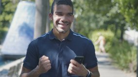 Medium shot of excited young Caucasian man in dark blue T-shirt texting on phone outside, receiving good news, saluting. Lifestyle, communication concept