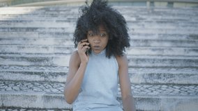 Pretty young Afro-american woman with curly hair and in jeans sleeveless dress sitting outside, talking on phone, looking upset and disappointed, gesticulating. Lifestyle, communication concept